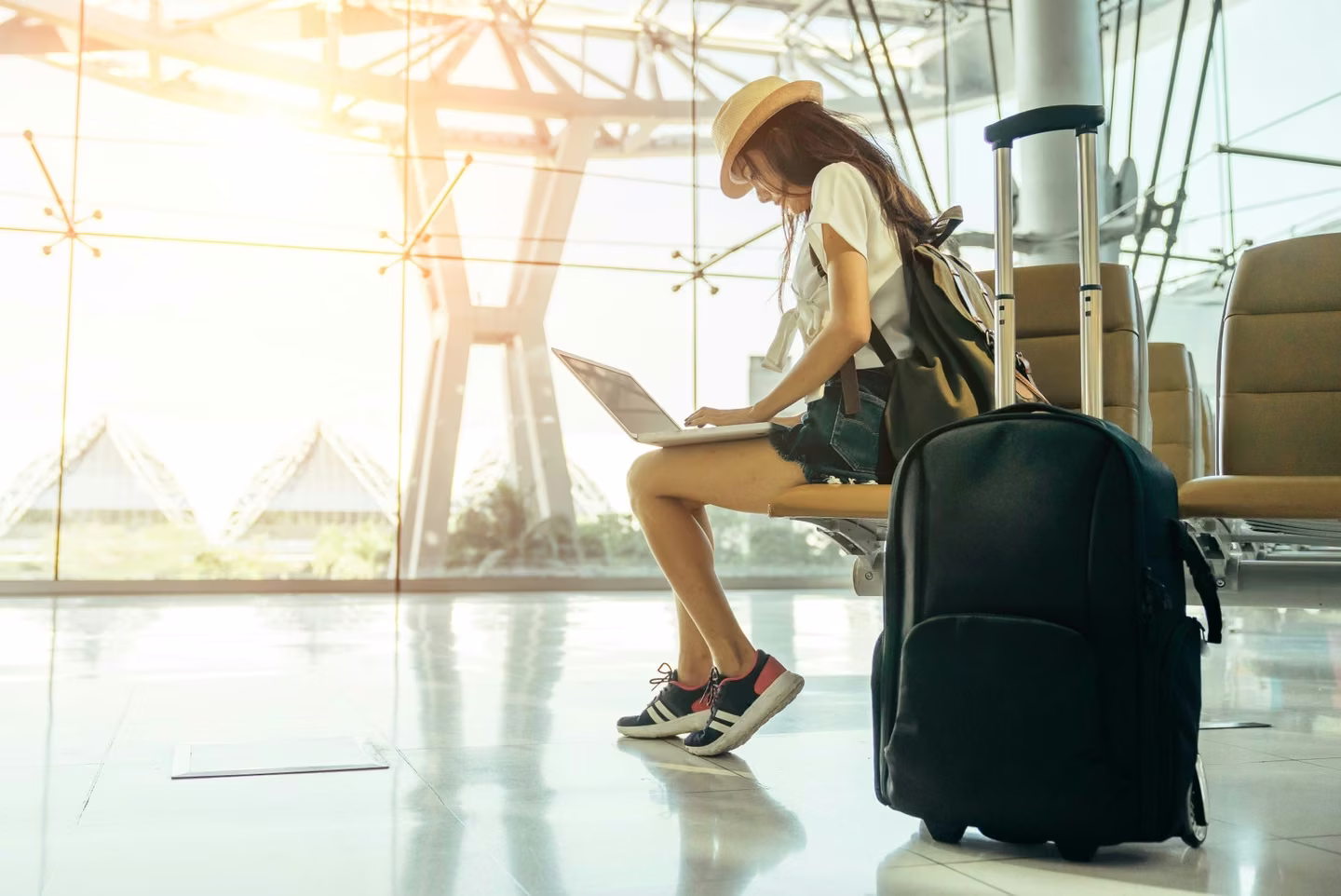 A person wearing a hat and casual clothes sits on a bench at an airport terminal, using a laptop. A wheeled suitcase is beside them. Sunlight streams through the large windows in the background, evoking thoughts of travel preparations and perhaps locating immediate care or urgent care facilities en route.
