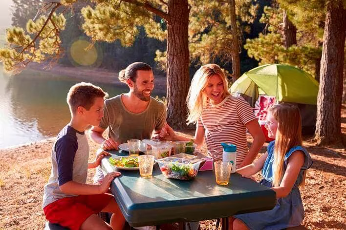 A family of four, including two adults and two children, enjoys a meal together at an outdoor table beside a lake, with a pitched tent and trees in the background, just a short distance from the walk-in clinic.