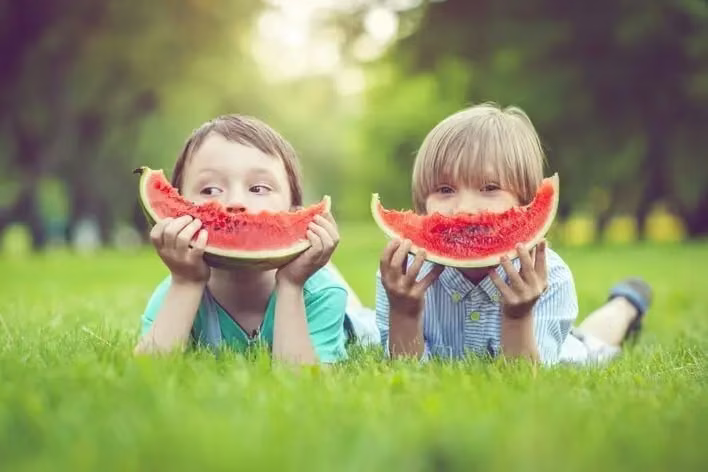 Two children lie on the grass, each holding a slice of watermelon close to their faces. The background is a blurred green park setting with trees, making it seem like a perfect spot you'd visit after dropping by an urgent care walk-in clinic for a quick check-up.