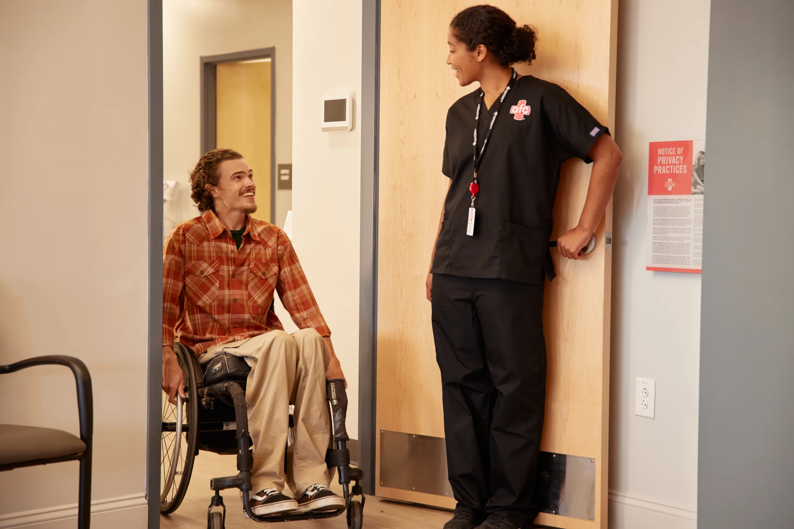 A man in a wheelchair wearing a plaid shirt and beige pants is smiling and conversing with a healthcare worker. The healthcare worker, dressed in black scrubs with a lanyard and ID badge, is standing in the doorway, smiling back at the man. A notice of privacy practices is visible on the wall beside them. The setting appears to be a medical facility or clinic.
