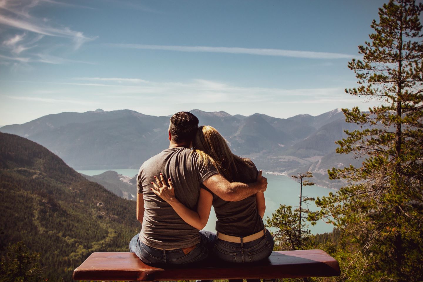 Two individuals sit on a bench overlooking a lush mountain landscape with a serene lake. They embrace, one’s arm around the other, gazing at the view.