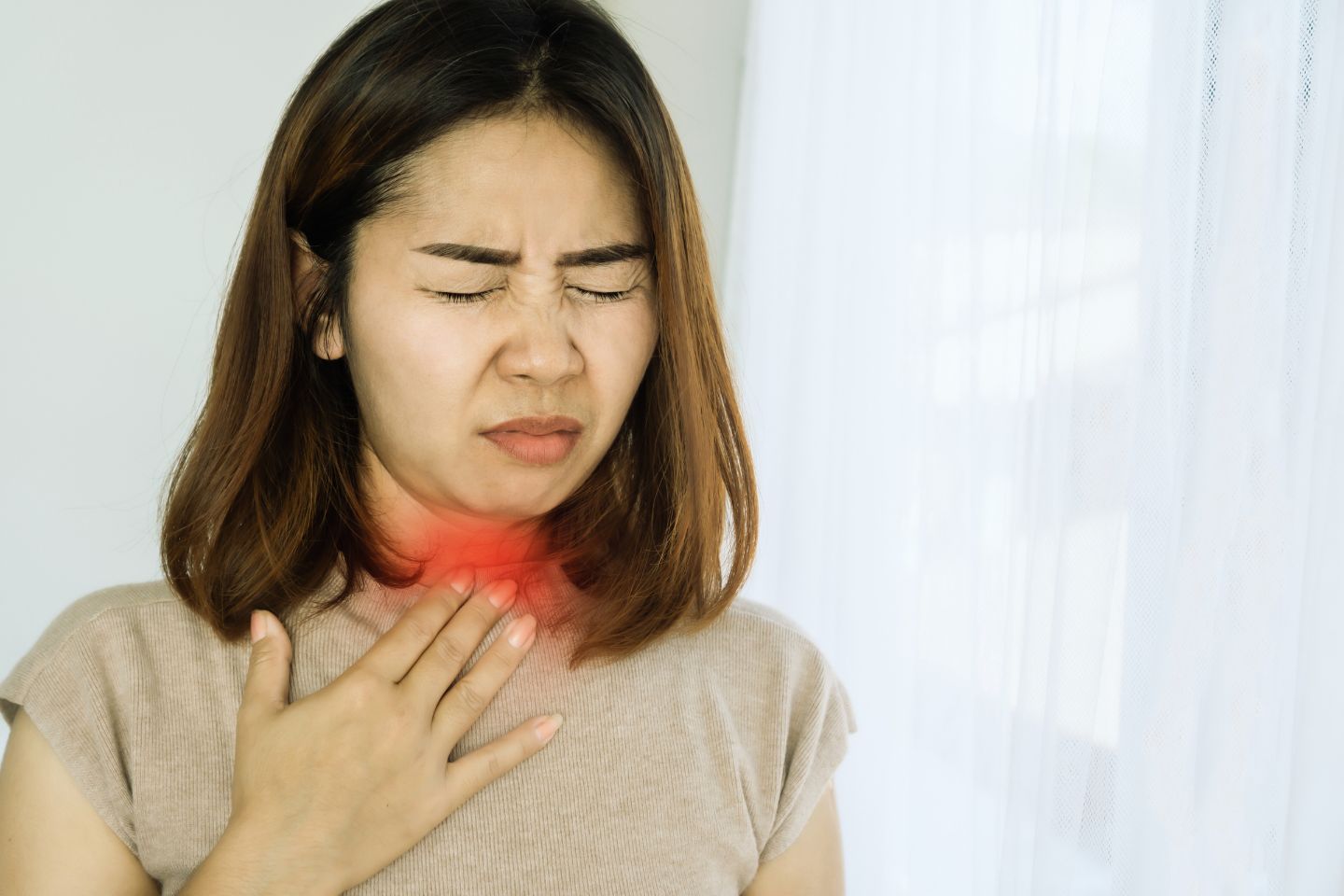 A woman experiencing discomfort or pain in her throat, which is highlighted with a red glow to indicate the affected area. She is grimacing and holding her throat, suggesting she might have a sore throat, possibly due to an infection such as strep throat.