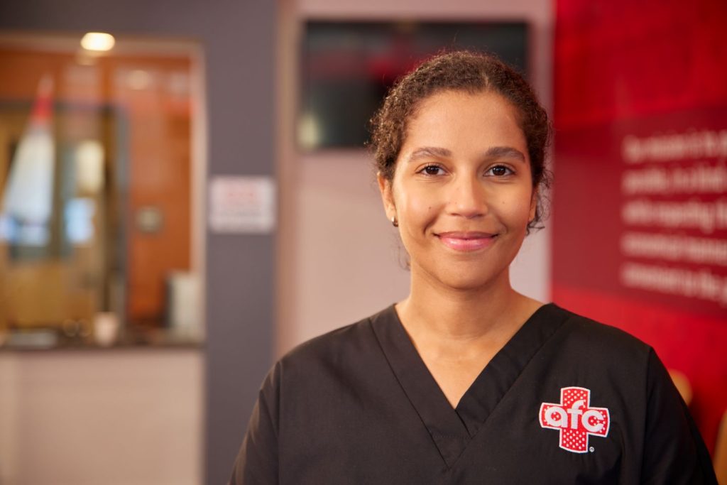 A young woman wearing black medical scrubs with a red and white logo stands indoors, smiling at the camera. The background is a blurred interior of a healthcare facility.
