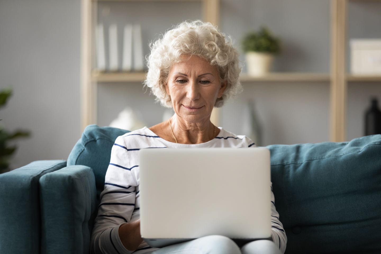 An elderly woman with curly gray hair is sitting on a teal sofa, using a laptop. She appears focused and content as she looks at the screen