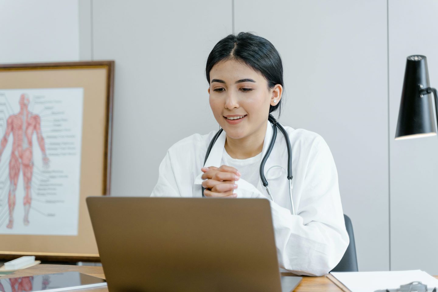A woman who appears to be a doctor or healthcare professional. She is wearing a white coat and a stethoscope around her neck. She is smiling and seems to be engaged in a conversation, likely through a video call on a laptop in front of her.
