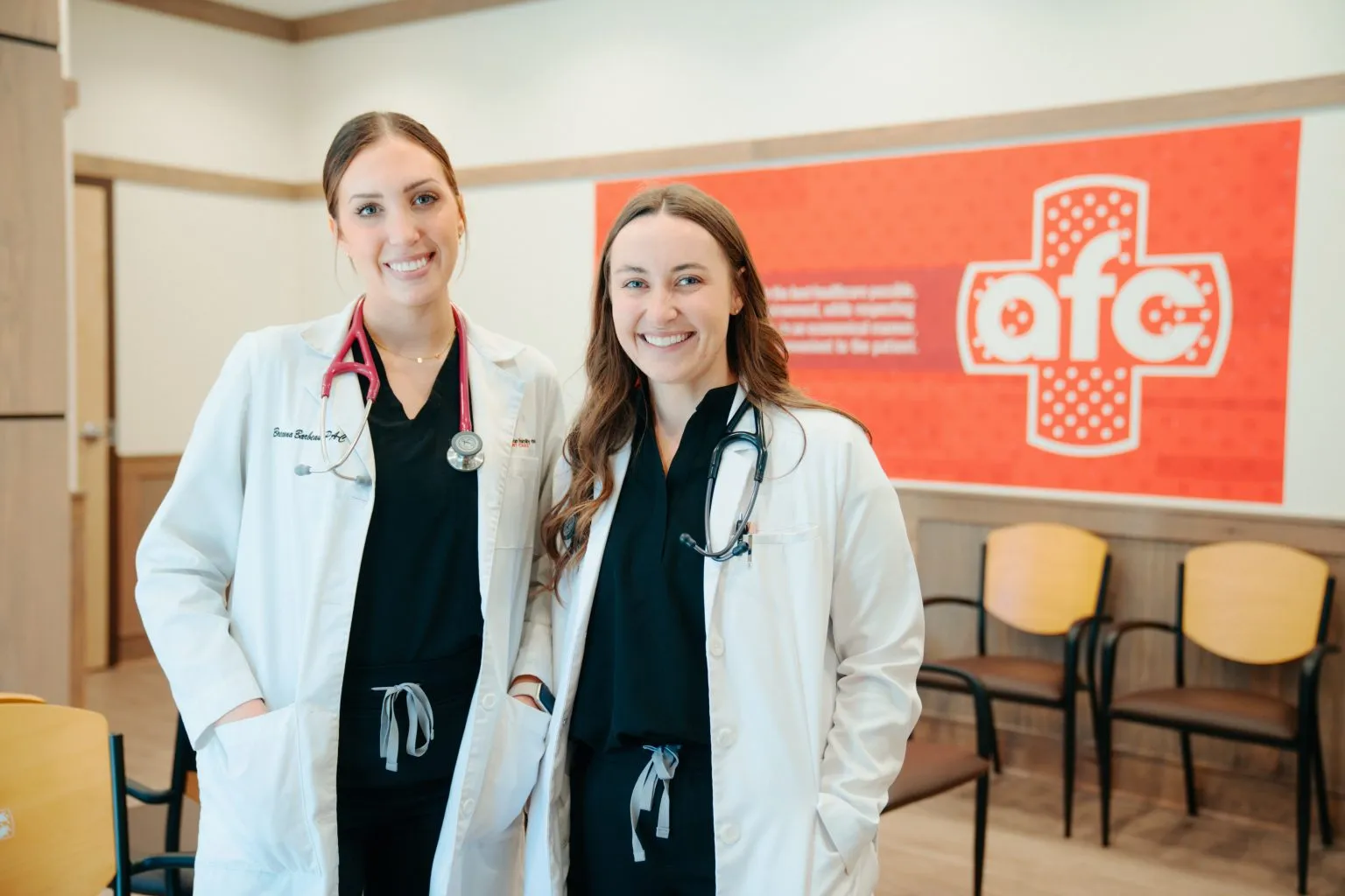 Two smiling women in medical coats stand in a room with chairs and a red healthcare logo in the background. Both have stethoscopes around their necks.