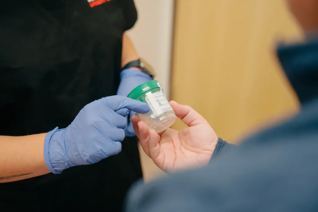 A person wearing blue gloves hands a clear container with a green lid to another person.